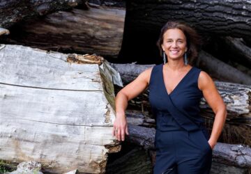 Lächelnde HR-Geschäftsführerin mit schulterlangen braunen Haaren trägt einen ärmellosen dunkelblauen Jumpsuit. Eine Hand ruht auf einem hellen, quer liegenden Baumstamm, dahinter gestapelte Stämme. Natürliche Lichtstimmung; Signal: Leadership & Bodenständigkeit.