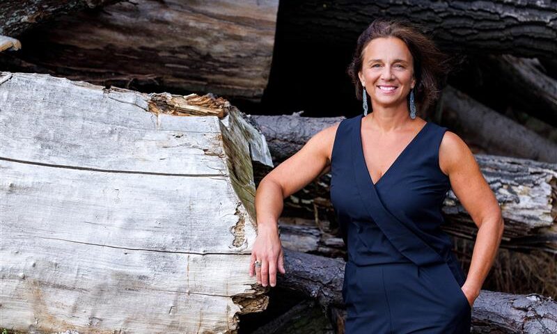Lächelnde HR-Geschäftsführerin mit schulterlangen braunen Haaren trägt einen ärmellosen dunkelblauen Jumpsuit. Eine Hand ruht auf einem hellen, quer liegenden Baumstamm, dahinter gestapelte Stämme. Natürliche Lichtstimmung; Signal: Leadership & Bodenständigkeit.