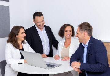 Vier Kolleg:innen im Meeting an rundem Tisch mit Laptop – Teamwork, Beratung und kollaboratives Arbeiten im modernen Büro.